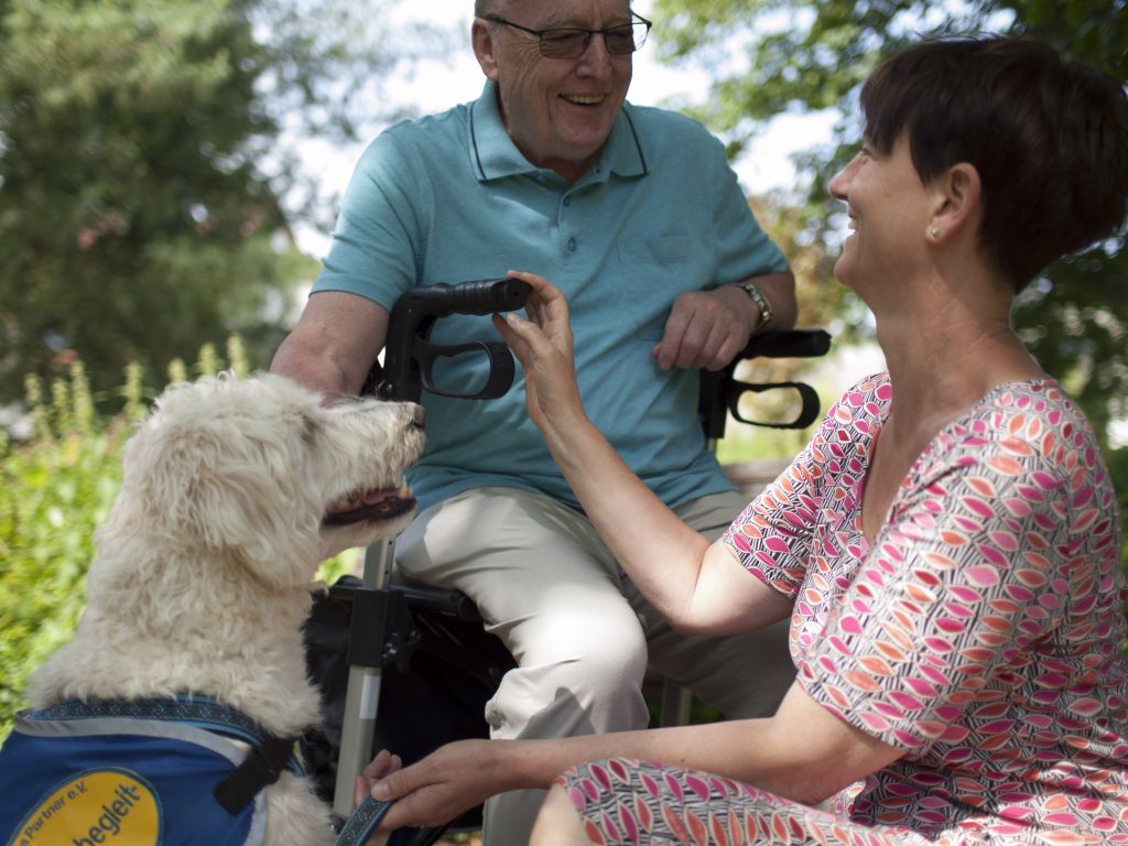 Ein fröhlicher älterer Mann sitzt auf einer Gehhilfe. Davor kniet eine Frau mit Pflege Job bei der kreuznacher diakonie neben einem Pflege Hund.