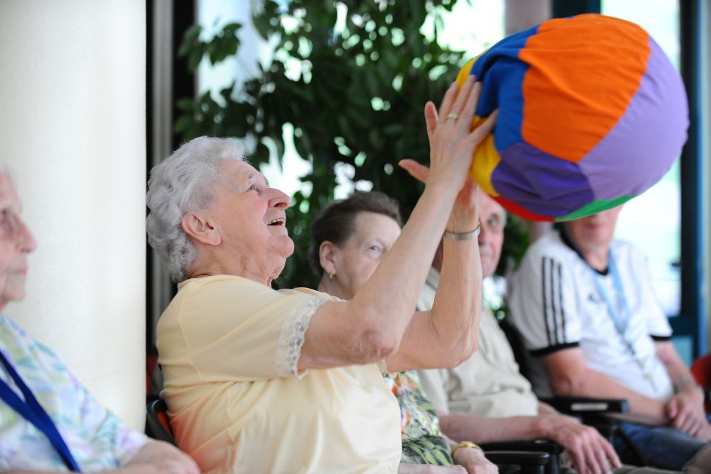 Mehrere Senioren bei der kreuznacher diakonie nebeneinander. Eine alte Dame fängt gerade einen bunten Ball auf.