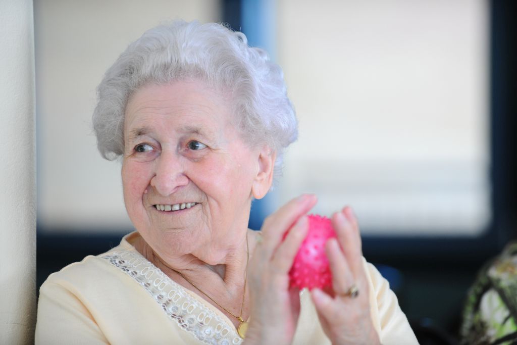 Eine fröhliche alte Dame bei der kreuznacher diakonie, die einen pinken Massageball in den Händen hält.
